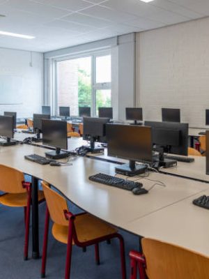 Computer education in classroom on high school. On this long table you see many desktop computers with keyboards and computer mouses. Also many chairs for the children to sit and work with the computer. In this classroom the students get computer lessons so they get prepared for our digital society as a lifestyle